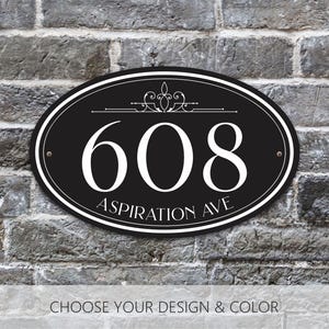 May include: An oval black house address plaque featuring the white number 608 and the text Aspiration Ave in a serif font. The sign includes a decorative flourish above the number, a double-line border, and two small mounting holes on the sides.