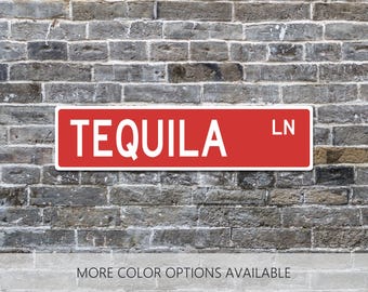 May include: A rectangular red street sign with a white border and white lettering that reads TEQUILA LN. The sign is mounted horizontally against a gray brick wall background. The text MORE COLOR OPTIONS AVAILABLE is printed at the bottom.