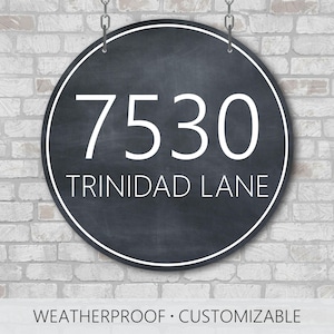 May include: A circular black sign with a white border and white text reading 7530 TRINIDAD LANE. The sign features a matte finish and is suspended by two metal chains at the top. The text is centered with the numbers above the street name.