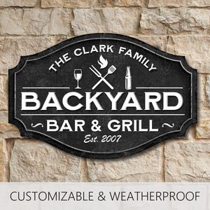 May include: Black and white sign with the text "The Clark Family Backyard Bar & Grill Est. 2007". The sign has a graphic of a wine glass, a bottle of beer, and crossed grilling tools.
