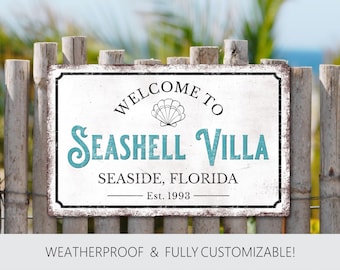 Könnte beinhalten: Ein rechteckiges Schild mit dem Text "WELCOME TO SEASHELL VILLA SEASIDE, FLORIDA Est. 1993" in schwarzer und teal-farbener Schrift. Das Schild zeigt eine Muschel-Grafik und ist an einem Holzzaun befestigt. Der Text "WEATHERPROOF & FULLY CUSTOMIZABLE!" steht unten.