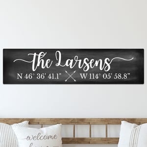 May include: A rectangular black sign with a chalkboard-style finish featuring the white text "the Larsens" in a cursive script, followed by the coordinates "N 46° 36' 41.1" W 114° 05' 58.8"" and a small graphic of two crossed arrows.