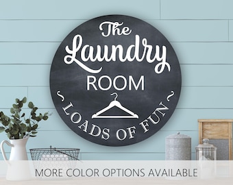May include: A circular black sign with a chalkboard-style finish featuring white text that reads The Laundry Room and Loads of Fun, accompanied by a white graphic of a clothes hanger centered between the two phrases in a clean, bold font.