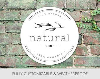 May include: A circular white sign featuring a black graphic of a leafy branch, the text "natural" in a serif font, and the text "SHOP" in a bold sans-serif font. The sign includes the text "100% NATURAL" and "100% ORGANIC" with circular border lines.