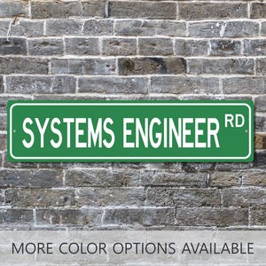 May include: A green rectangular street sign with the words "SYSTEMS ENGINEER RD" in white lettering. The sign is mounted on a weathered brick wall. Below the sign, the text "MORE COLOR OPTIONS AVAILABLE" is displayed.