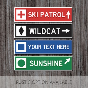 May include: Four rectangular signs stacked vertically. The top is red with a white cross and 'SKI PATROL' with an arrow. The second is black with a diamond and 'WILDCAT' with an arrow. The third is blue with a square and 'YOUR TEXT HERE'. The bottom is green with a circle and 'SUNSHINE' with an arrow.