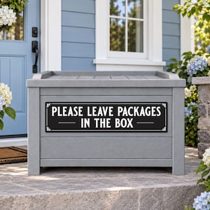 May include: A rectangular gray storage box featuring a black horizontal sign attached to the front. The sign has white text that reads "PLEASE LEAVE PACKAGES IN THE BOX" with decorative flourishes on the left and right sides of the text.
