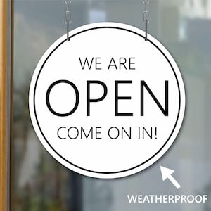 May include: A white round sign with black text that reads "We are open come on in!" hanging from a chain on a glass door. The sign is weatherproof.