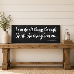 May include: A rectangular black sign with white cursive text that reads, I can do all things through Christ who strengthens me. - Philippians 4:13. The sign has a matte finish and is positioned horizontally on a wooden bench surface.