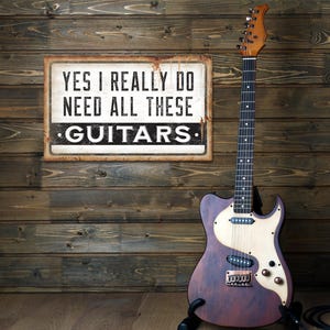May include: A vintage-style sign with the text "YES I REALLY DO NEED ALL THESE GUITARS" hangs on a wooden wall. A brown and cream electric guitar stands in front of the wall, with a black cable on the floor.