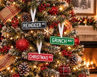 Peut inclure: Trois décorations suspendues rectangulaires en forme de panneaux de rue, chacune avec une boucle en ruban blanc. Un panneau noir indique REINDEER RD, un vert GRINCH WAY et un rouge CHRISTMAS RD, le tout sur un sapin de Noël décoré.