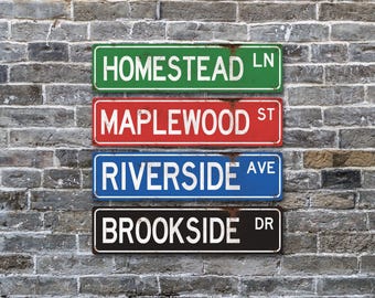 May include: Four rectangular metal street signs stacked vertically. The top sign is green with white text reading HOMESTEAD LN. The second is red with white text reading MAPLEWOOD ST. The third is blue with white text reading RIVERSIDE AVE. The bottom is black with white text reading BROOKSIDE DR.