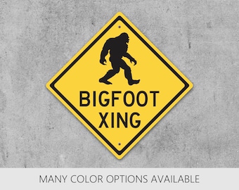 May include: A diamond-shaped yellow warning sign with a black border and black text that reads BIGFOOT XING. The sign features a black silhouette graphic of a Bigfoot creature walking toward the right side of the sign.
