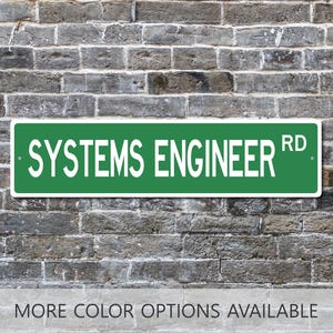 May include: A rectangular green street sign with white text that reads SYSTEMS ENGINEER RD. The sign features a white border and two small mounting holes on the left and right sides. The text is in a bold, sans-serif, all-caps font style.