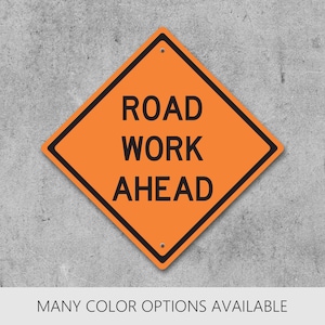 May include: A diamond-shaped orange road sign with a black border and the black text ROAD WORK AHEAD. The sign features two small circular holes at the top and bottom center points, indicating mounting points for installation on a post or wall.