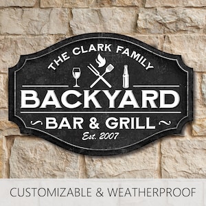 May include: A black, oval-shaped sign with white text and graphics. The sign reads THE CLARK FAMILY BACKYARD BAR & GRILL Est. 2007. Graphics include a wine glass, crossed grilling utensils with a flame, and a bottle. The sign has a textured finish.