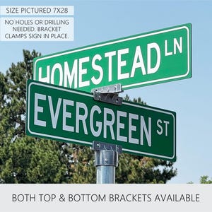 May include: Green street signs with white lettering. The top sign reads "HOMESTEAD LN" and the bottom sign reads "EVERGREEN ST".  The signs are mounted on a metal pole with brackets.  The text "SIZE PICTURED 7X28" is visible at the top of the image.  The text "NO HOLES OR DRILLING NEEDED. BRACKET CLAMPS SIGN IN PLACE." is also visible.