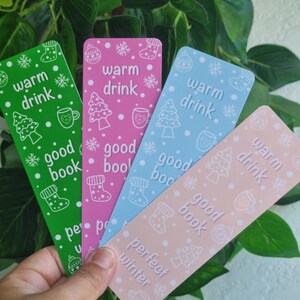 Könnte beinhalten: Vier farbige Lesezeichen mit Winterthema. Die Lesezeichen zeigen ein weißes Schneeflockenmuster mit dem Text "warm drink", "good book" und "perfect winter".
