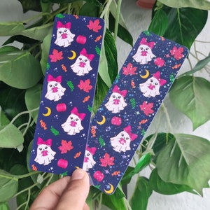 May include: Two blue and black bookmark designs featuring a white ghost with a pink bow holding a red apple. The background is a dark blue with stars, candy corn, and autumn leaves.