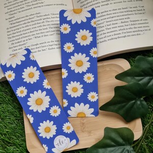 May include: Two blue bookmarks featuring a white daisy pattern. The bookmarks are displayed on a wooden surface with green grass in the background. The text on the bookmark reads "Free Cake for Everyone".