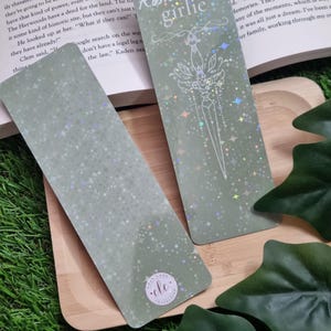 May include: Three holographic bookmarks with a green background and a white design. The bookmarks feature a dagger, stars, and the text "Romantasy Girlie".