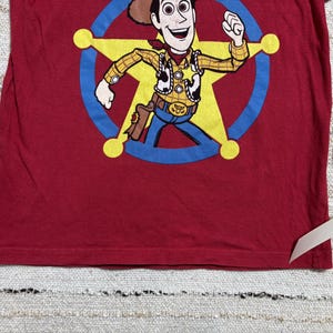 May include: A long-sleeved, red t-shirt featuring a cartoon image of Woody from Toy Story. Woody is depicted in his sheriff attire, inside a yellow star with a blue outline. The shirt is made of a soft material.