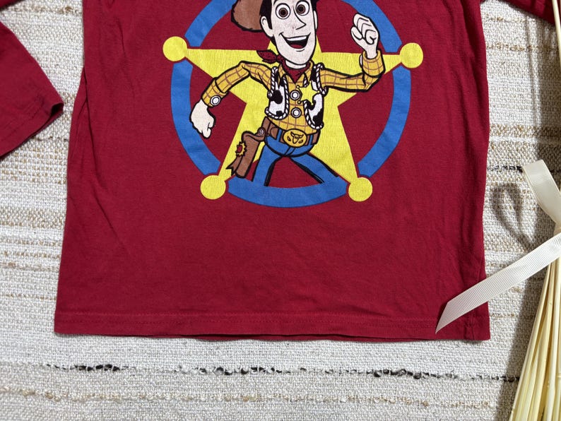 May include: A long-sleeved, red t-shirt featuring a cartoon image of Woody from Toy Story. Woody is depicted in his sheriff attire, inside a yellow star with a blue outline. The shirt is made of a soft material.