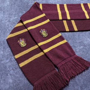 Peut inclure: Écharpe à rayures bordeaux et dorées avec une bordure à franges. L'écharpe présente un écusson sur chaque extrémité. Fabriquée dans une matière douce, elle est idéale pour l'hiver.