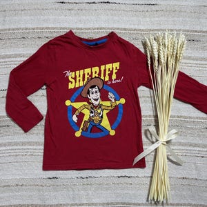 May include: A long-sleeved, red t-shirt featuring a cartoon image of Sheriff Woody from Toy Story. The graphic includes the text "The Sheriff is here!" in yellow and white. A bundle of wheat tied with a white ribbon is also present.