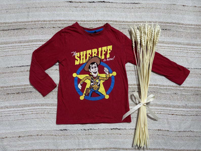 May include: A long-sleeved, red t-shirt featuring a cartoon image of Sheriff Woody from Toy Story. The graphic includes the text "The Sheriff is here!" in yellow and white. A bundle of wheat tied with a white ribbon is also present.