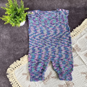 May include: A knitted baby romper in shades of blue, purple, and green. The romper has a sleeveless design with a round neckline and is adorned with small pink buttons on the shoulders. The garment is made of a soft, textured yarn.