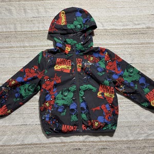 Op de afbeelding: Zwarte jas met capuchon met Marvel Comics-personages zoals Spider-Man en de Hulk. De jas heeft een ritssluiting over de hele lengte en het Marvel Comics-logo in rood en geel. De jas is ontworpen voor kinderen.