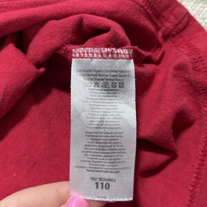 May include: Close-up of a red garment with a white tag attached. The tag displays care instructions and the number "110". The fabric is folded, revealing the texture and stitching details. The garment appears to be made of a soft material.