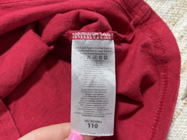 May include: Close-up of a red garment with a white tag attached. The tag displays care instructions and the number "110". The fabric is folded, revealing the texture and stitching details. The garment appears to be made of a soft material.