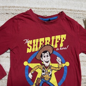 May include: A maroon long-sleeved shirt featuring a graphic of Woody from Toy Story. The design includes the text "The Sheriff is here!" in yellow, with Woody in a sheriff's outfit inside a blue star.
