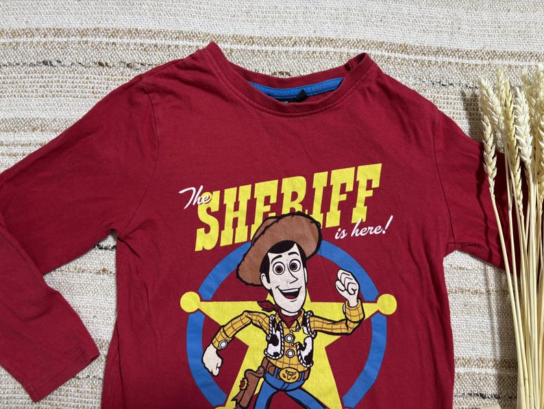 May include: A maroon long-sleeved shirt featuring a graphic of Woody from Toy Story. The design includes the text "The Sheriff is here!" in yellow, with Woody in a sheriff's outfit inside a blue star.