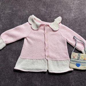May include: A pink and white knitted baby cardigan with a scalloped collar and cuffs. The cardigan has a button-up front and is paired with a small, matching handbag with a blue heart detail. The set is designed for a baby or toddler.