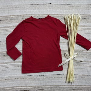May include: A long-sleeved, burgundy-colored shirt is displayed on a textured, neutral-toned surface. A bundle of dried wheat stalks, tied with a white ribbon, rests beside the shirt. The shirt is the focal point, showcasing its solid color and simple design.
