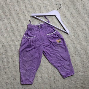 May include: A pair of lavender-colored children's pants with suspenders, featuring two front pockets with white and lavender trim. A small embroidered patch is visible on one leg. The pants are hanging on a white wooden hanger.