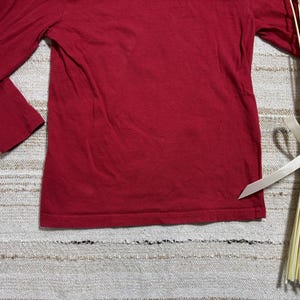 May include: A solid red long-sleeve shirt. The shirt is made of a soft material and has a crew neck. The sleeves extend to the wrists. The shirt is displayed on a textured surface with a bundle of dried stems tied with a white ribbon.