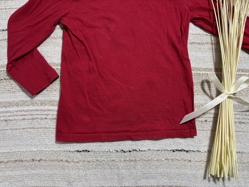 May include: A solid red long-sleeve shirt. The shirt is made of a soft material and has a crew neck. The sleeves extend to the wrists. The shirt is displayed on a textured surface with a bundle of dried stems tied with a white ribbon.