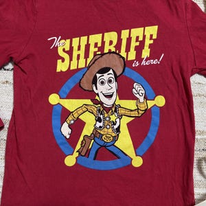 May include: A long-sleeved, red t-shirt featuring a cartoon image of Woody from Toy Story. The graphic includes Woody in a sheriff's outfit inside a star, with the text "The Sheriff is here!" in yellow and white.