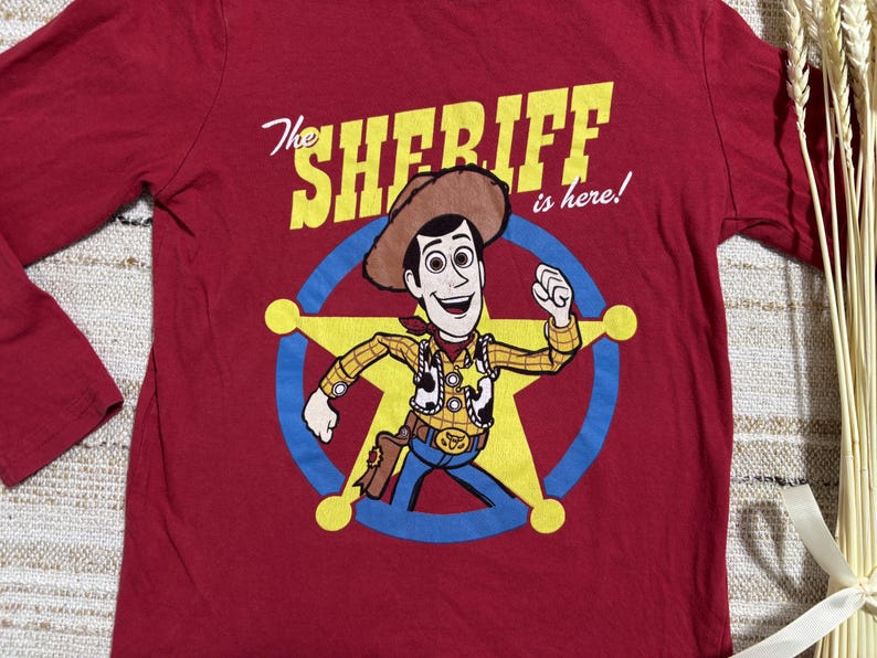 May include: A long-sleeved, red t-shirt featuring a cartoon image of Woody from Toy Story. The graphic includes Woody in a sheriff's outfit inside a star, with the text "The Sheriff is here!" in yellow and white.