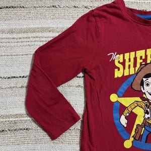 May include: A long-sleeved, red t-shirt featuring a cartoon illustration of Woody from Toy Story. The graphic includes Woody in a sheriff's outfit, a sheriff's badge, and the text "The Sheriff is here".