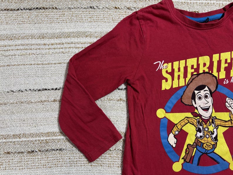 May include: A long-sleeved, red t-shirt featuring a cartoon illustration of Woody from Toy Story. The graphic includes Woody in a sheriff's outfit, a sheriff's badge, and the text "The Sheriff is here".