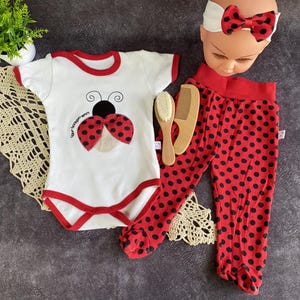 May include: A baby outfit set. The set includes a white bodysuit with a ladybug design, red trim, and matching red polka dot trousers. A wooden comb and a headband with a bow are also included. Designed for infants.
