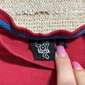 May include: Close-up of a red t-shirt collar with a blue trim. A black tag displays the "Toy Story 4" logo and the number "110". The shirt is made of a soft material.
