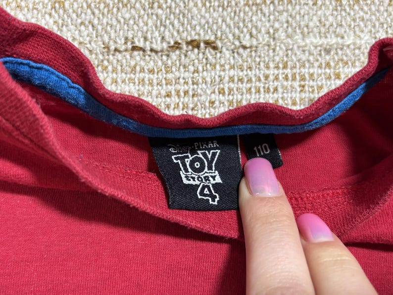 May include: Close-up of a red t-shirt collar with a blue trim. A black tag displays the "Toy Story 4" logo and the number "110". The shirt is made of a soft material.
