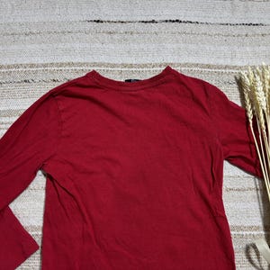 May include: A long-sleeved, solid red shirt. The shirt is made of a soft material and has a crew neck. A bundle of wheat stalks tied with a white ribbon is on the right side of the shirt.
