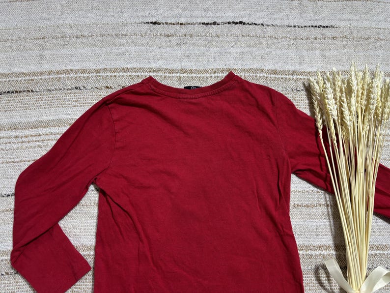 May include: A long-sleeved, solid red shirt. The shirt is made of a soft material and has a crew neck. A bundle of wheat stalks tied with a white ribbon is on the right side of the shirt.